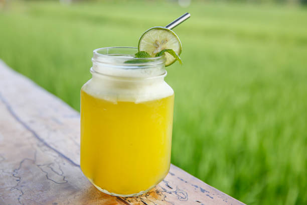 Immunity-Boosting Citrus Juice Close-up shot of fresh orange lemonade juice on a glass jar with a slice of lime on top and a reusable metal straw. The drinking glass is on a wooden table with a background of a lush green paddy field. mason jar lemonade stock pictures, royalty-free photos & images