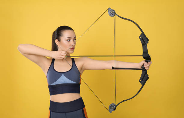 sportliche junge frau üben bogenschießen auf gelbem hintergrund - archery bow arrow women stock-fotos und bilder