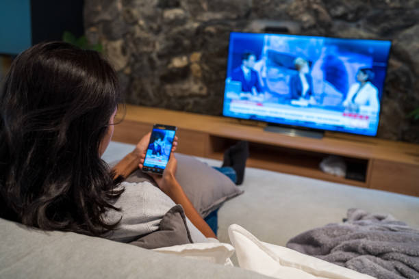 Young woman watching news on television and smart phone Rear view of young woman watching news on television and smart phone while sitting on sofa in living room. tv stock pictures, royalty-free photos & images