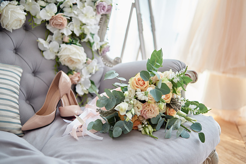 Bridal bouquet of roses, freesia, brunia and eucalyptus with satin ribbons and beige bride shoes on high heels on chesterfield sofa, copy space. Wedding morning preparations