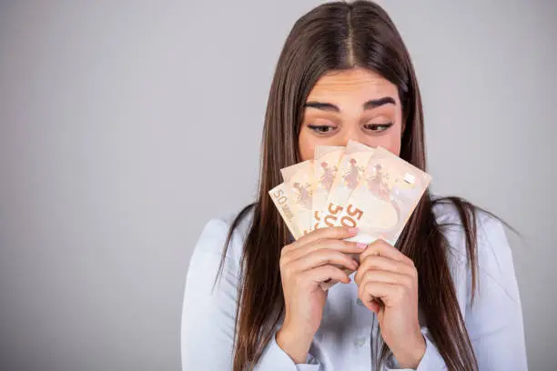 Concept shot of an ecstatic young woman holding a fanned-out wad of Euros in her hand. Close up portrait of shocked surprised girl peeking out fan of much money with wide open eyes hiding her half face isolated on gray background