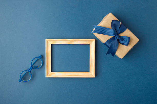 istock Father's day composition. Gift box with blue ribbon, eyeglasses and frame mock up on blue background. Top view flat lay with free space for text. Greeting card for dad, copy space, invitation 1311251739