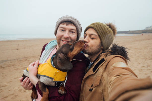 Puppy Kisses LGBTQI couple and their dachshund dog walking on the beach in Tynemouth in the North East of England. They are taking a selfie on one of their mobile phones. man gay stock pictures, royalty-free photos & images