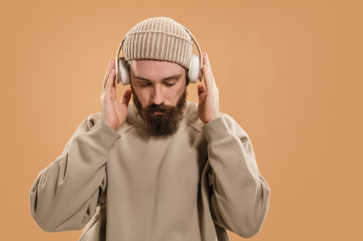 Listening to music. Half-length portrait of man in headphones and hat isolated over light yellow background. Mustache and beard. Concept of human emotions, facial expression, fashion, culture.