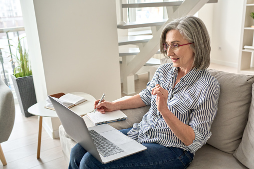Mature adult old professional woman watching web training, online webinar on laptop computer remote working or e learning class from home office, video calling having virtual meeting, writing notes.