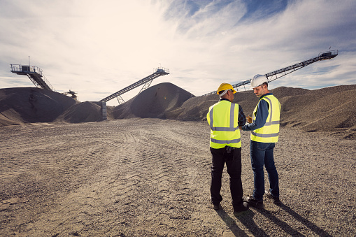 Open-pit mine engineer and worker wearing protective clothes and helmets standing in front of pile of gravel, discussing plans. Back view, wide angle view.