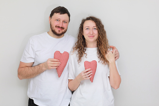 smiling man and woman in white t-shirts with red hearts in their hands lovingly looking at each other. couple in love on a white background