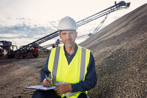 Open-pit mine engineer wearing protective clothes and white helmet squatting next to pile of gravel, holding clipboard and looking at camera.
