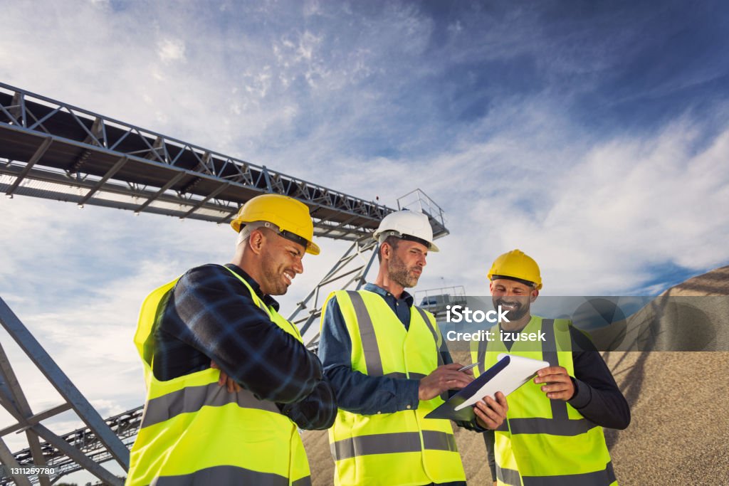 Open-pit mine workers Open-pit mine engineer and workers wearing protective clothes and helmets standing in front of pile of gravel, discussing plans. Occupation Stock Photo