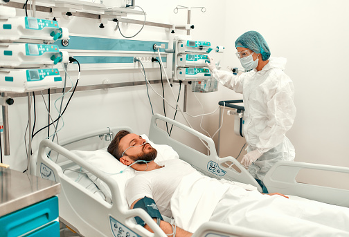 A doctor in a protective suit checks a sick man for the coronavirus disease covid-19, who is in an intensive care unit in a modern hospital.