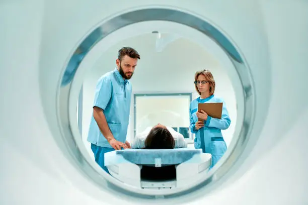 The patient lies on CT or MRI, the bed is moved inside the machine, scanning her body and brain under the supervision of a doctor and a radiologist. In a medical laboratory with high-tech equipment.