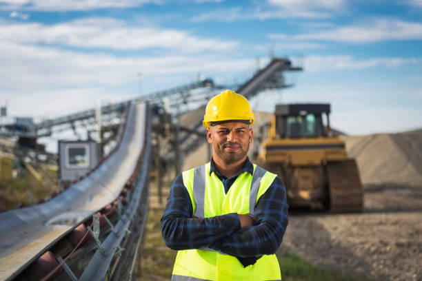 Portrait of open-pit mine worker Open-pit mine worker wearing protective clothes and yellow helmet standing next to conveyor belt with arms crossed, smiling at camera. construction worker stock pictures, royalty-free photos & images