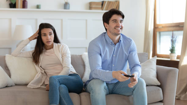 улыбающийся мужчина смотрит телевизор, скучает разгневавшаяся женщина, сидящая отдельно - family television fighting watching стоковые фото и изображения