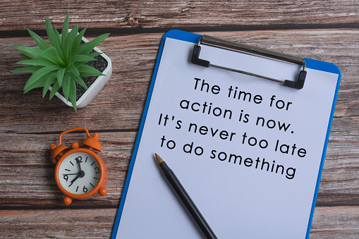Text on blue clipboard with pen, alarm clock and potted plant on wooden desk - The time for action is now, it is never too late to do something