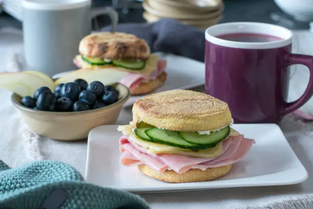 toasted breakfast sandwich with ham, cheese, and cucumber served with a bowl of fresh fruits and a mug of tea on a kitchen table at home.