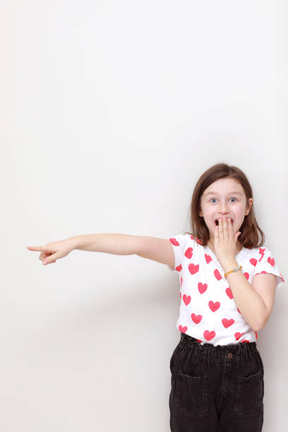 Cute smiling kid girl in white t-shirt with red hearts, stylish black jeans wonderingly pointing her finger looking to the side on white background. valentines day Cute smiling kid girl in white t-shirt with red hearts, stylish black jeans wonderingly pointing her finger looking to the side on white background. valentines day. wonderingly stock pictures, royalty-free photos & images