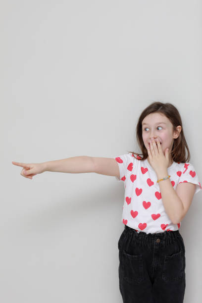 Cute smiling kid girl in white t-shirt with red hearts, stylish black jeans wonderingly pointing her finger looking to the side on white background. valentines day Cute smiling kid girl in white t-shirt with red hearts, stylish black jeans wonderingly pointing her finger looking to the side on white background. valentines day. wonderingly stock pictures, royalty-free photos & images