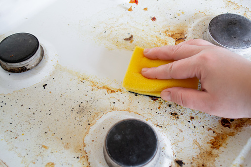 A hand with a yellow wash sponge washes the very dirty greasy surface of the gas stove. After the sponge, a clean trace remains.