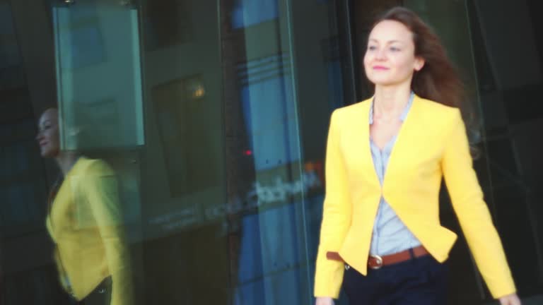 Focus on the reflection. Slow motion. Young business woman in yellow jacket walking near office building in the big city.