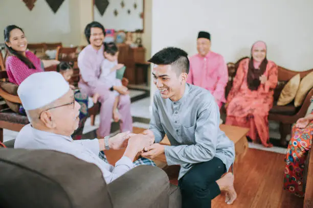 malay muslim grandson in traditional clothing showing apologize gesture to his grandfather during Aidilfitri celebration  malay family at home celebrating hari raya