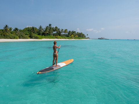 She stands on paddle board and enjoys relaxation on tropical turquoise lagoon in the Maldives