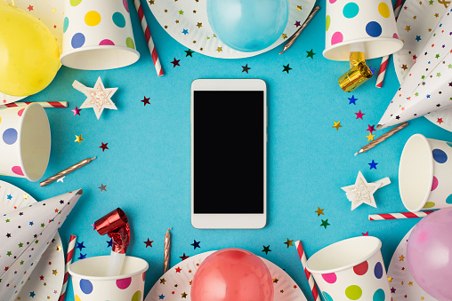 Top view photo of birthday table composition party accessories and smartphone with copyspace in the middle on isolated blue background