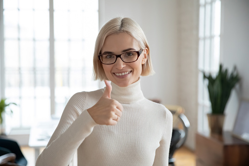 Headshot portrait of smiling young Caucasian woman in glasses show thumb up give recommendation. Happy businesswoman recommend good company service or office employment. Success concept.