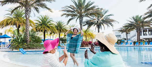 A multi-ethnic group of three senior women in their 60s on vacation. Two of them are sitting on the pool deck watching their Hispanic friend showing off her new swimsuit and beach coverup and sun hat.