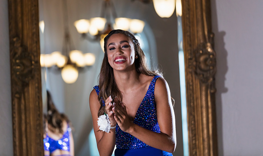 A mixed race teenage girl, 16 years old, standing in a restaurant by a mirror, wearing a prom dress and wrist corsage.