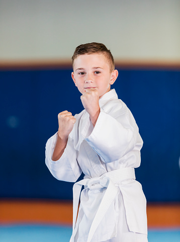A boy in a gym learning taekwondo. He is looking at the camera, fists raised in a pose, wearing a dobok with a white belt. He is 7 years old, mixed race Hispanic and Caucasian.
