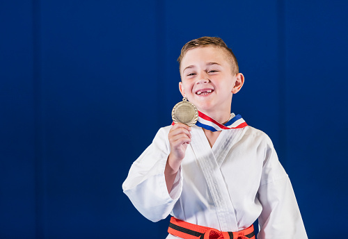 A proud boy in a taekwondo uniform, a dobok, shows off his medal for winning a competition. He is 7 years old, mixed race Hispanic and Caucasian.