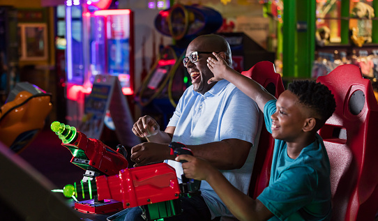 An African-American father and son having fun at an amusement arcade, playing a game pointing toys laser guns at a screen, playfully competing. The main focus is on the man, in his 50s, laughing as the 11 year old boy tries to push him to keep him from winning.
