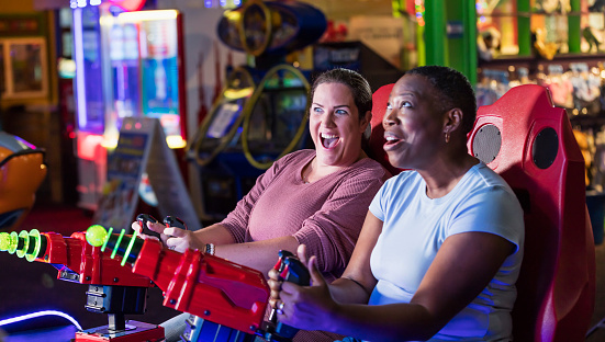 Two women having fun at an amusement arcade. They are sitting side by side playing a game, pointing toys laser guns at a screen. The focus is on the Caucasian woman, in her 30s. Her African-American friend is in her 40s. They are both laughing.