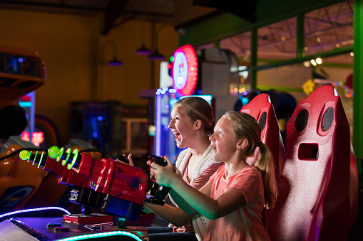Two sisters having fun at an amusement arcade, playing a game pointing toys laser guns at a screen, competing against each other. The focus is on the 14 year old teenage girl. Her little sister is 6.