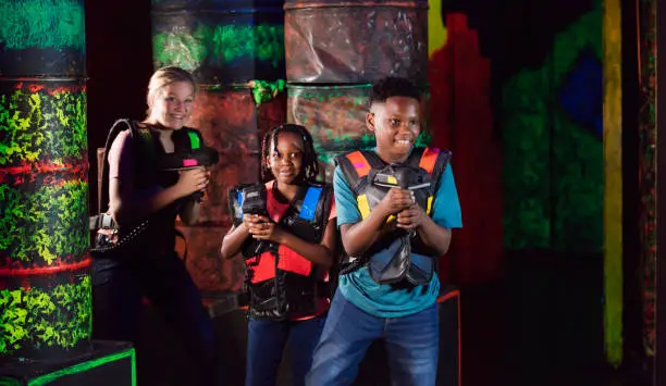 A multi-ethnic group of three children playing laser tag at an amusement arcade. They are walking, looking around for targets. The main focus is on the 11 year old African-American boy and his 7 year old sister.