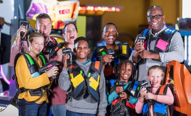 A multi-ethnic group of children and adults, friends and family, having fun together at an amusement arcade. They are dressed for a game of laser tag, holding their toy laser guns, smiling at the camera. There are two families. The parents are in their 30s to 50s. The African-American boy is 11 years old, his sister is 7, and the Caucasian girls are 14 and 6.