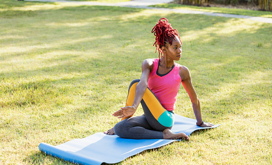 A mid adult African-American woman in her 30s practicing yoga outdoors, on an exercise mat on the grass. She is doing a half spinal twist.