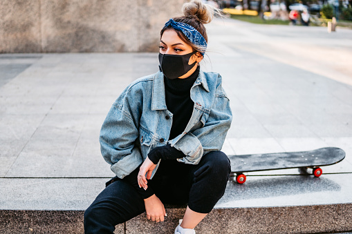 Young beautiful woman with skateboard wearing protective face mask in public park.