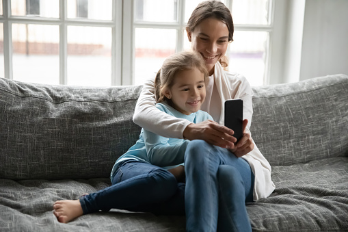 Close up happy mother and little daughter having fun with phone together, sitting on cozy couch at home, smiling mum hugging adorable child cute girl, making video call to relatives, browsing apps