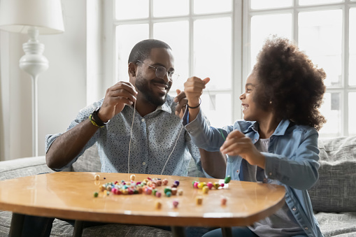 Excited girl and happy dad stringing colorful wooden beads together. Young father helping kid to train craft skills, enjoying creative handmade activity with daughter. Family leisure time concept