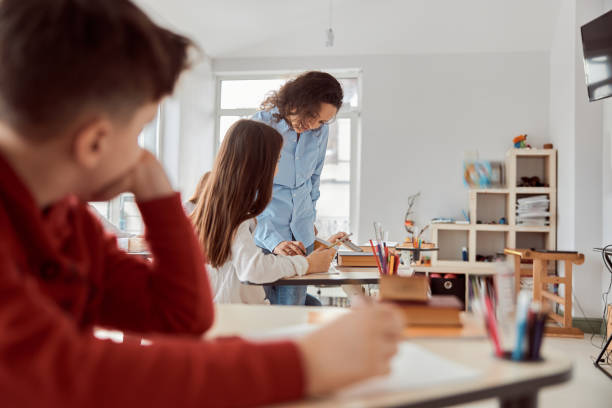 istock Young teacher helps to read her student. Elementary school kids sitting on desks and reading books in classroom 1311335832
