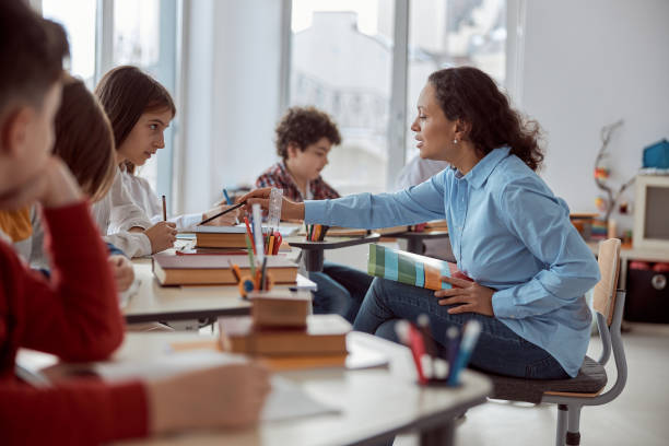 istock Young teacher helps to read her student. Elementary school kids sitting on desks and reading books in classroom 1311335840