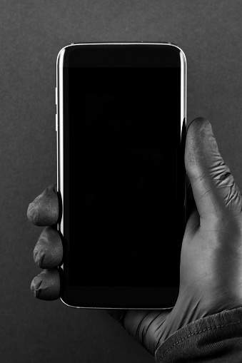 Hand in black protective glove holding smartphone on dark grey background - screen is ready for your message
