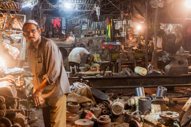 Mumbai, India. March 17th, 2014: A view of a street shop in Mumbai with a makeshift shelter made from tarpaulin and wood. Bicycles are parked in front of the shop, some with old carts attached, and the structure shows signs of wear. A sign written in Hindi and the Indian date format marks a point of interest on the building. In the background, modern apartment buildings contrast with the humble street scene. The image captures the juxtaposition of Mumbai's urban growth and traditional, simple livelihoods.