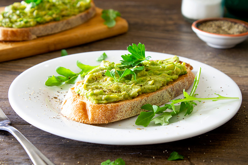 Fresh avocado toast on a white plate on a rustic wooden table. A chopping board with more in the background. 
The avocado is sprinkled with chopped parsley and black pepper, and served with arugula. Stock photo.