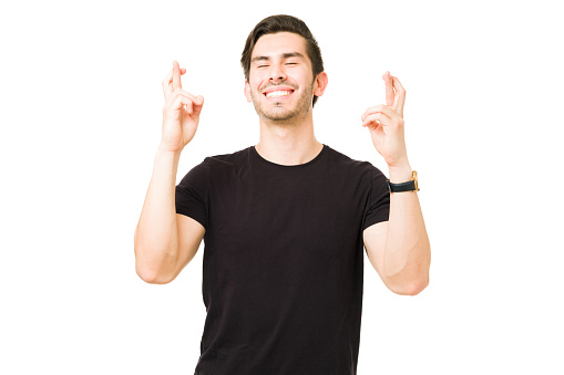 Handsome young man wishing his dreams come true with his fingers crossed. Latin man in his 20s feeling lucky against a white background