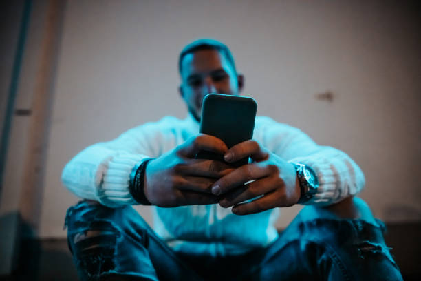 Young man checking news and posts on mobile phone in the city at night Young man sitting on urban stairs checking news and social media posts on his smart phone in llluminated city street at night. Shot from below, selective focus on smart phone, while reading news and posts on his mobile phone. Real People Millennial Generation Modern Technology Urban Lifestyle Portrait. mobile phone lights up stock pictures, royalty-free photos & images