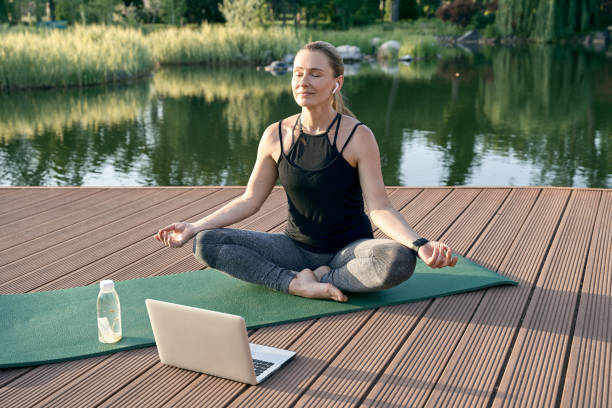 istock Serenity. Sporty relaxed woman meditating, sitting in Lotus pose while watching tutorial on a laptop, doing yoga on a mat in nature on a sunny day outdoors 1311350119
