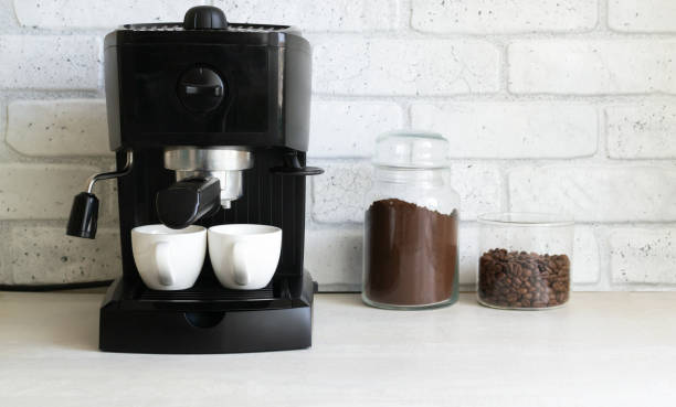 A black coffee machine is on the kitchen table, next to it are two glass jars with ground coffee and beans. stock photo