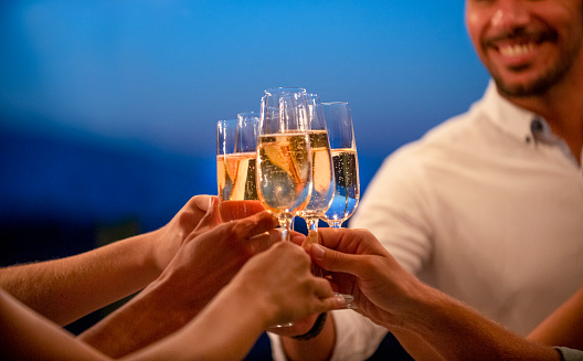 Group of friends toasting with champagne at night. They are standing outdoors in a restaurant at dusk. A man is visible smiling in the background. Close up of hands and champagne flutes